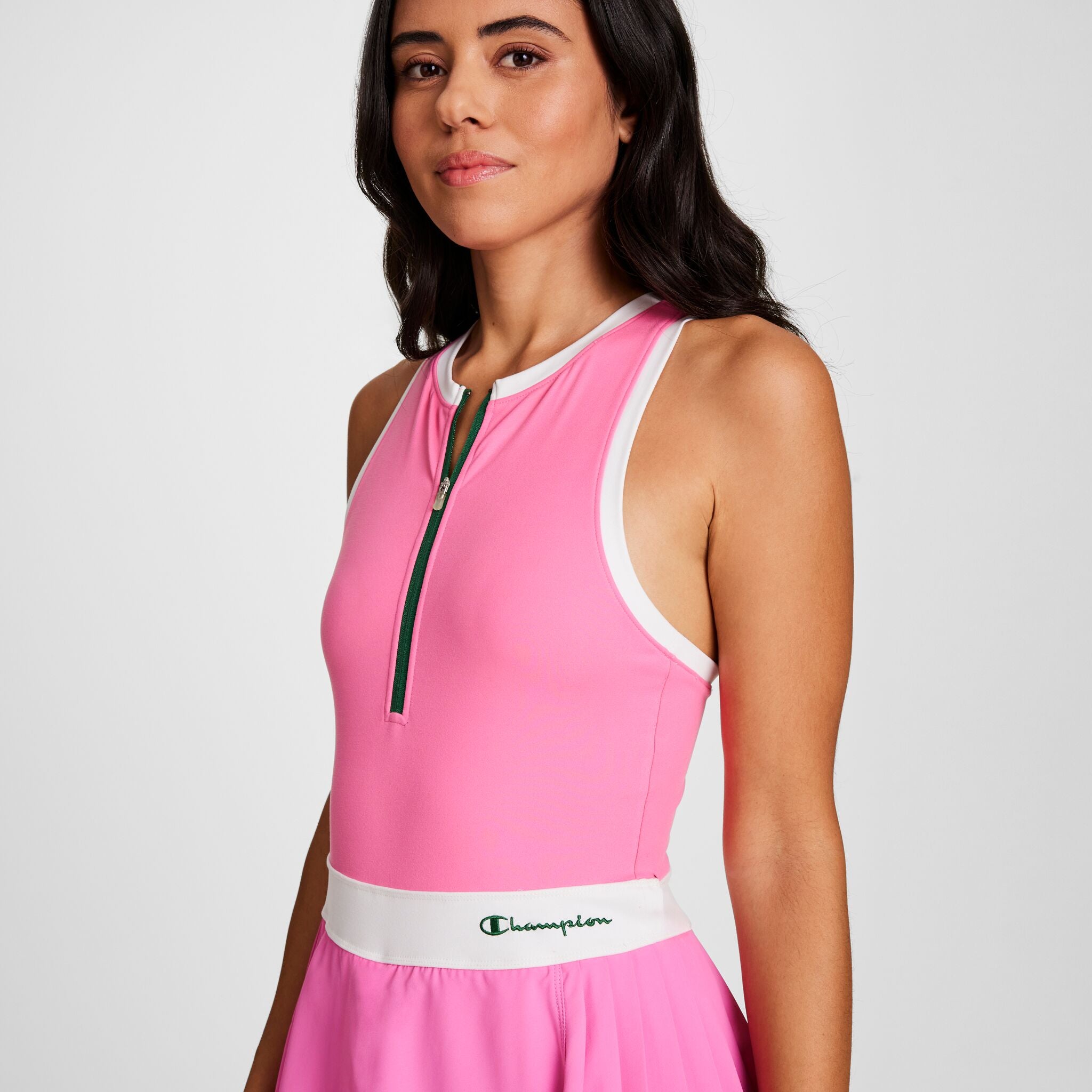 A woman with long dark hair wears the Champion Match Point Tennis Dress, featuring a sleeveless pink design with white trim, front zipper, embroidered script logo on the waistband, built-in shorts, and performance fabric, against a plain background.