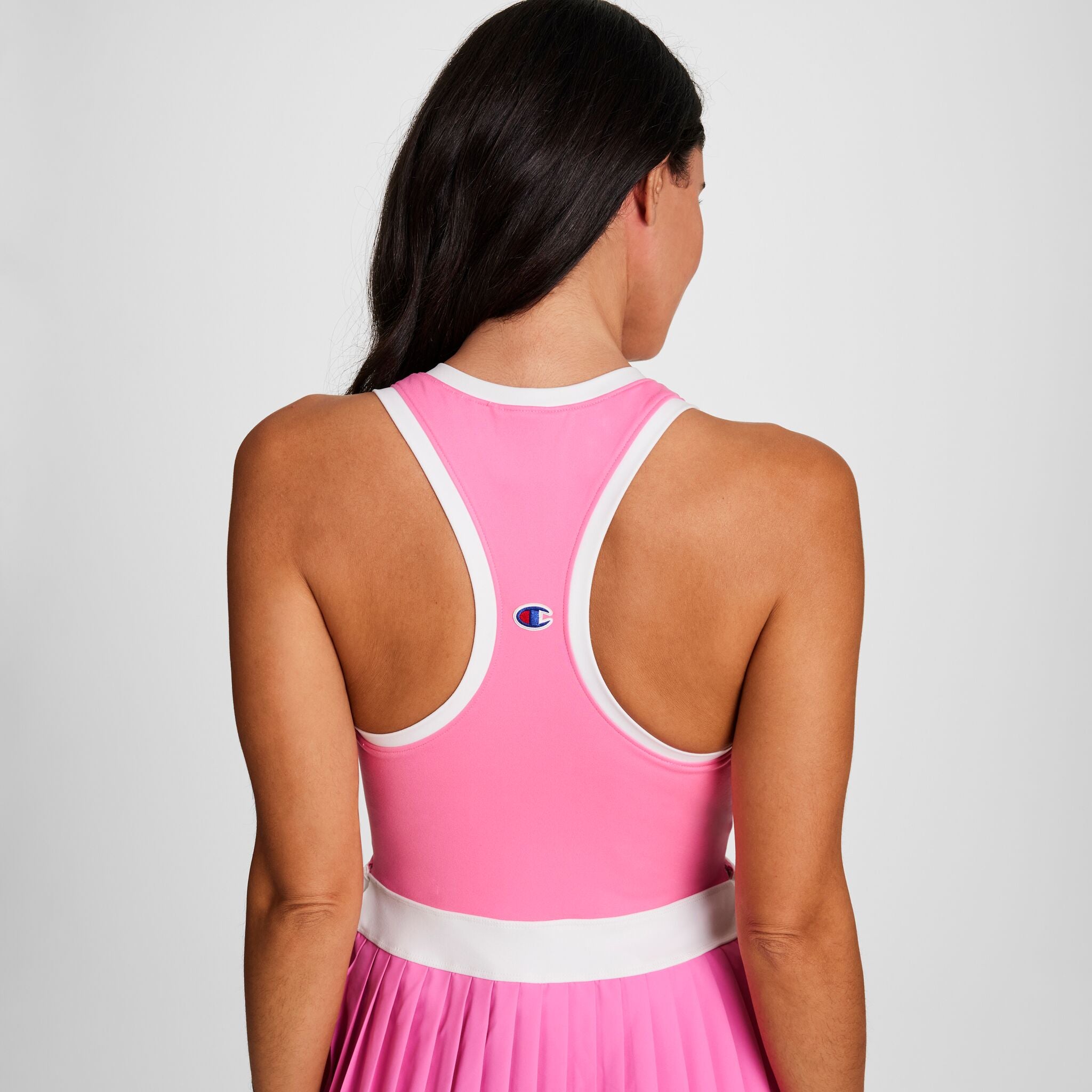 A woman with long dark hair faces away, wearing the Champion Match Point Tennis Dress featuring white trim, racerback style, pleated skirt, built-in inner short for comfort and coverage, and an embroidered script logo.