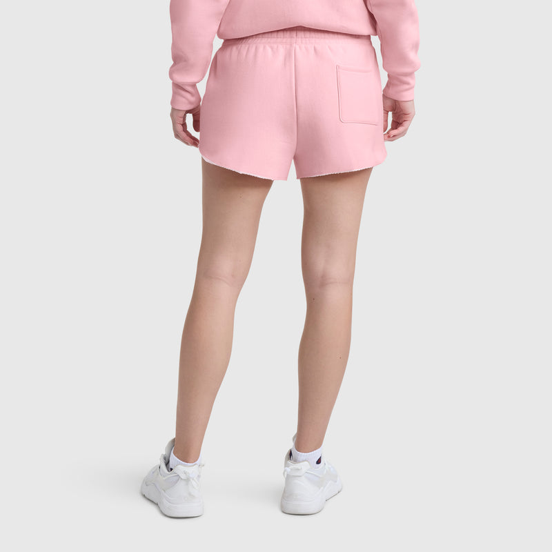 A person faces away wearing Champion 2.5" Swing Shorts (C Logo) in light pink with an elastic waistband and single back pocket, matching pink sweatshirt, and white sneakers against a plain light gray background.