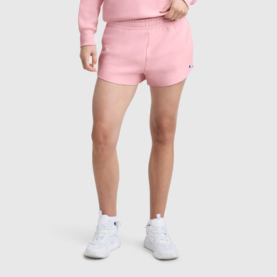 A person stands against a light gray background wearing a light pink sweatshirt, Champion 2.5" Swing Short with C Logo, and white sneakers. Their face is not visible.