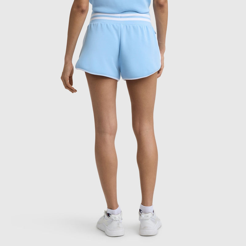 A person stands with their back to the camera, wearing Champion’s 3" Tennis Court Shorts with an embroidered script logo, white waistband and trim, paired with white sneakers against a plain light background.