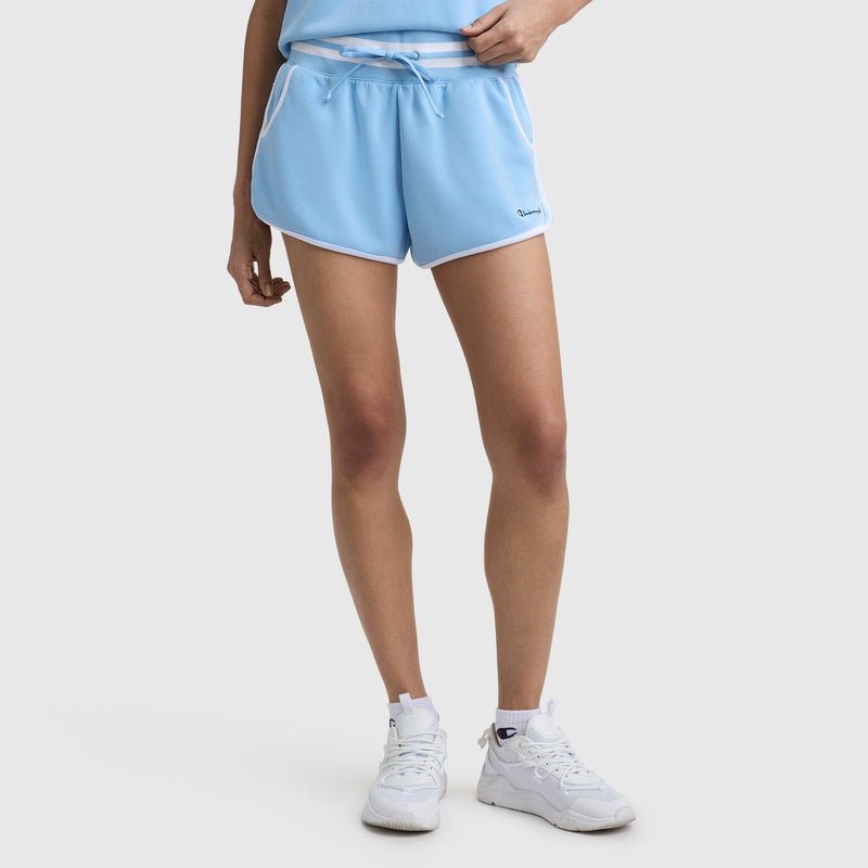 Model wearing Champion 3" Tennis Court Short with embroidered script logo in light blue with white trim, paired with a matching top and white sneakers. The photo shows only the lower torso and legs against a plain white background.