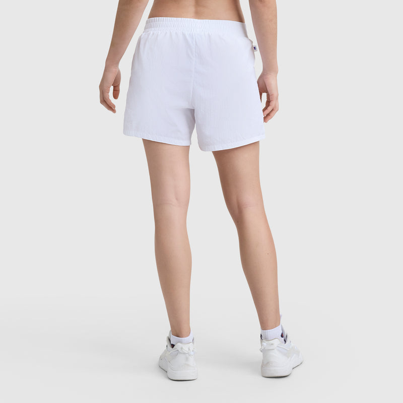 A person stands facing away, showing only their lower back and legs, wearing Champion 5" Courtside Short, C Logo, in white crinkle nylon with white sneakers against a plain light background.