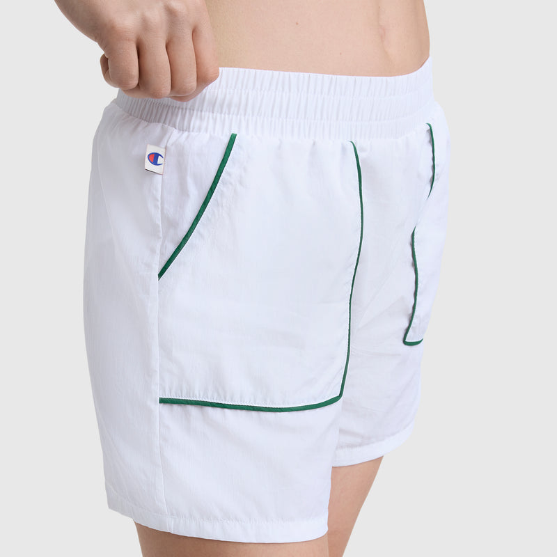 A person wears Champion's 5" Courtside Short in white crinkle nylon, featuring green pocket piping, an elastic waistband, and a small C logo patch on the left. The upper torso is not visible.