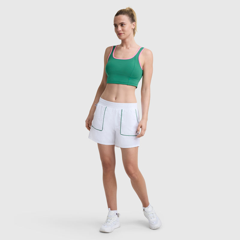 A woman stands against a light gray background, smiling slightly with her hair in a ponytail. She wears a green sports bra with pink trim and Champion’s 5" Courtside Short, C Logo, which features large front pockets, paired with white sneakers.