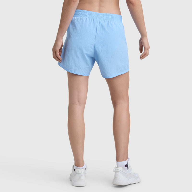 A person faces away, showing only their lower back and legs, wearing Champion 5" Courtside Short with C Logo in light blue, paired with white ankle socks and white sneakers against a plain light background.