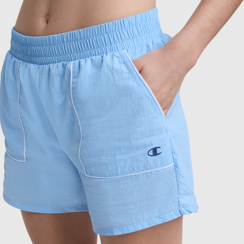Wearing Champion 5" Courtside Short with C Logo, these light blue nylon athletic shorts feature white trim and an embroidered logo on the left pocket, epitomizing classic sport heritage style with one hand in a pocket.