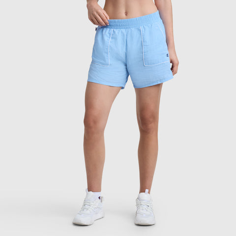 Someone wearing the Champion 5" Courtside Short, C Logo in light blue with white trim and white sneakers stands against a plain white background. Only their legs and part of their torso are visible.