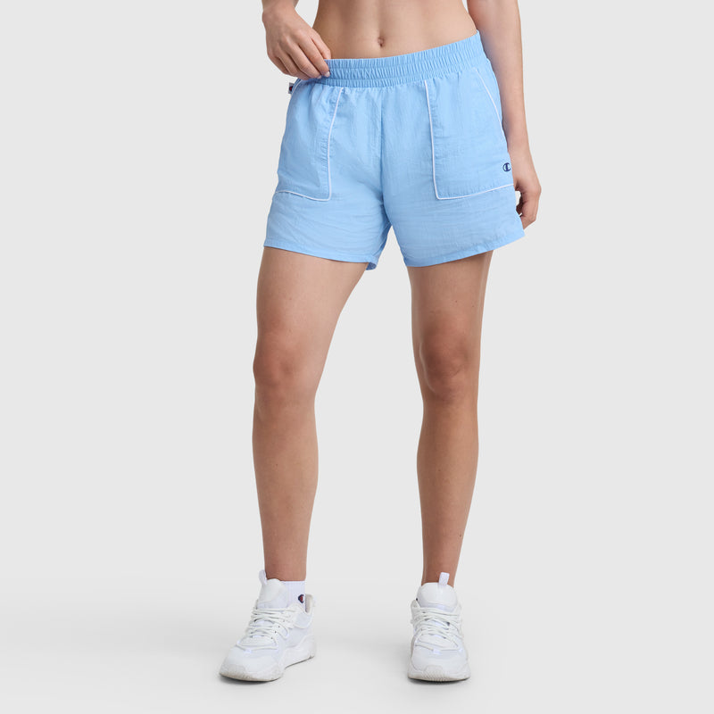 Someone wearing the Champion 5" Courtside Short, C Logo in light blue with white trim and white sneakers stands against a plain white background. Only their legs and part of their torso are visible.