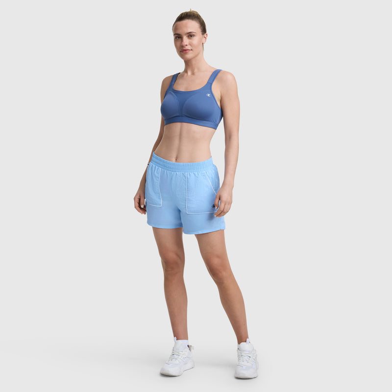 A woman faces forward in a blue sports bra and Champion 5" Courtside Short with C Logo—lightweight nylon athletic shorts with pockets—and white sneakers. Her pulled-back hair and the plain background evoke classic sport heritage style.