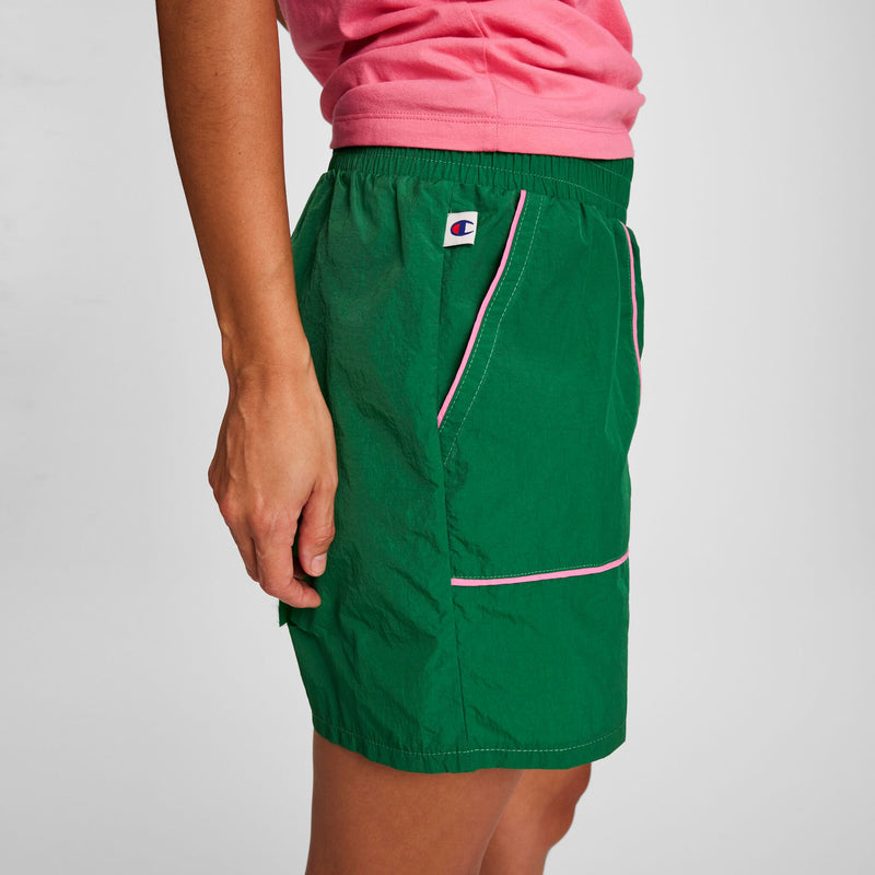 A person wearing a pink shirt and Champion’s 5" Courtside Short with C Logo in green and pink trim stands sideways, highlighting the crinkle nylon waistband. The background is plain and light-colored.