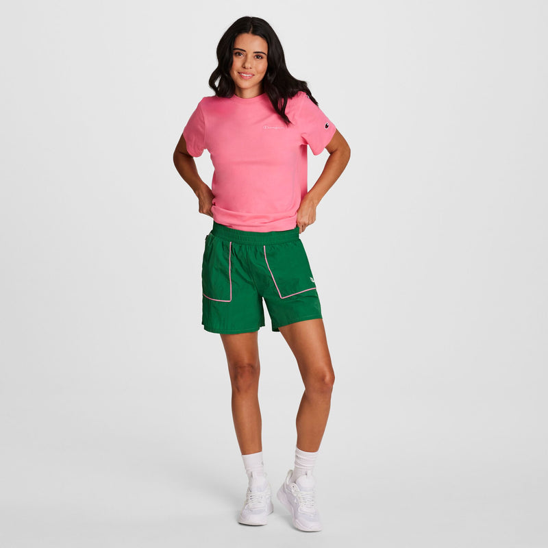 A woman with long dark hair stands confidently against a white background, wearing Champion's 5" Courtside Short with C Logo in green and a pink tee, styled with white socks and sneakers, hands on her hips and smiling.