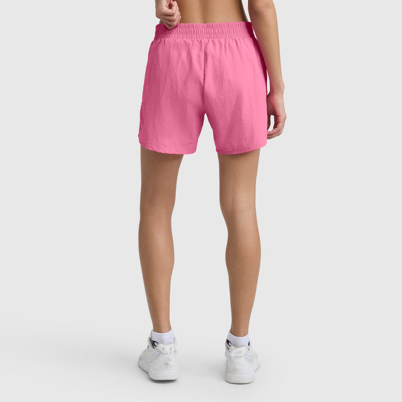 A person stands facing away, wearing bright pink Champion 5" Courtside Short with C Logo, white sneakers, and a white sports bra against a plain light background.