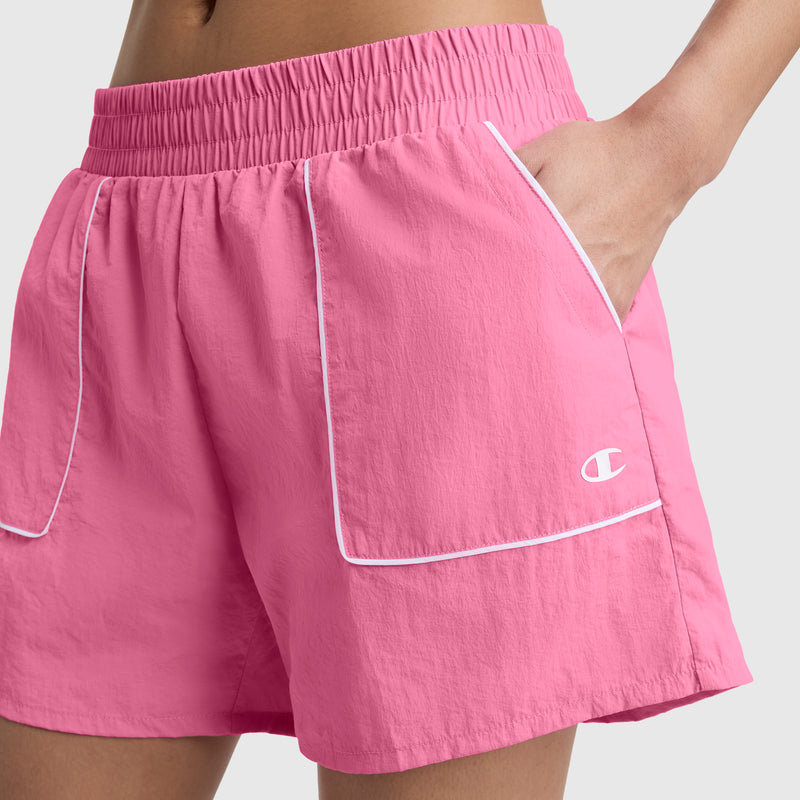 A person wearing Champion 5" Courtside Shorts in bright pink, featuring white piping, an elastic waistband, and a small white C logo on the left, with one hand in the pocket.
