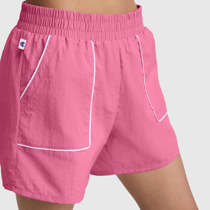 A person wears bright pink Champion 5" Courtside Short athletic shorts with white piping, an elastic waistband, and a small C logo on the left. The crinkle nylon shorts are set against a plain, light-colored background.
