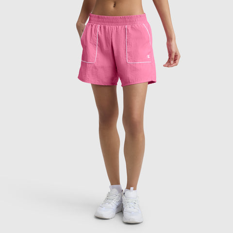 A person wears Champion's 5" Courtside Short, C Logo in bright pink with white trim and pockets, featuring an elastic waistband and loose fit, paired with white athletic shoes against a plain light background.