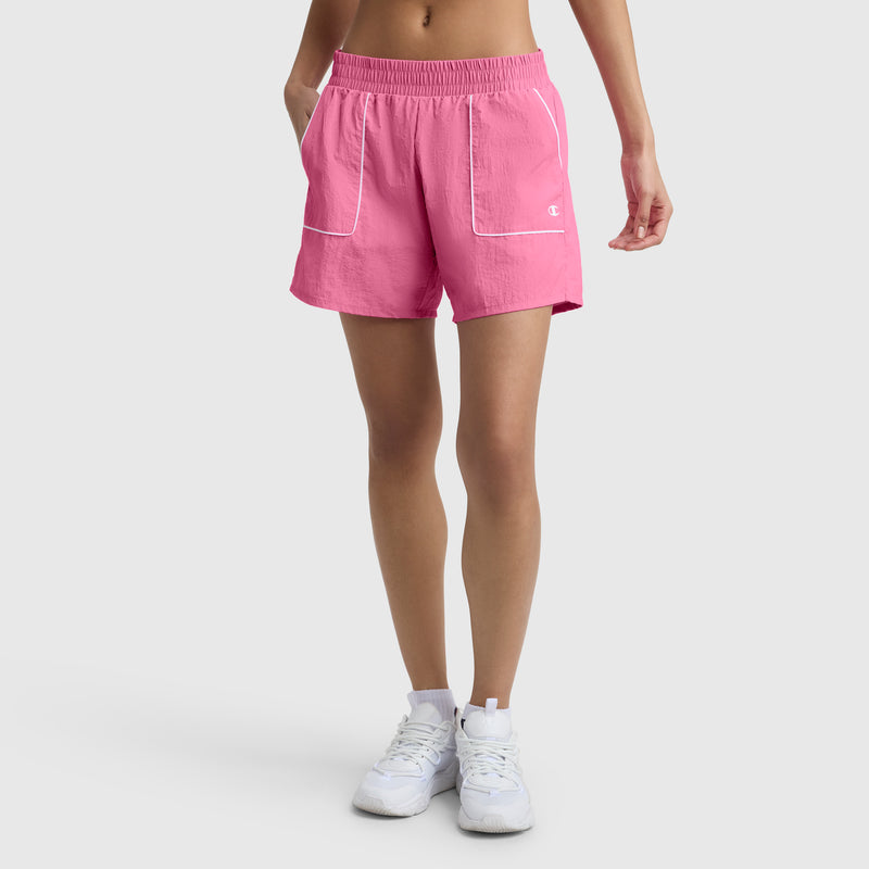 A person wears Champion's 5" Courtside Short, C Logo in bright pink with white trim and pockets, featuring an elastic waistband and loose fit, paired with white athletic shoes against a plain light background.