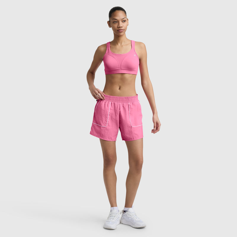 A woman poses against a plain background in a Champion 5" Courtside Short with C Logo, a pink sports bra, and white sneakers. Her hair is tied back and she stands casually with one hand by her side.