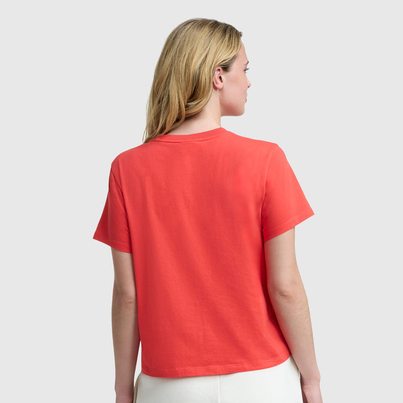 A person with blonde hair is shown from the back, wearing the Champion Modern Retro Baseball Graphic T-Shirt with a script logo and white pants against a plain light gray background.