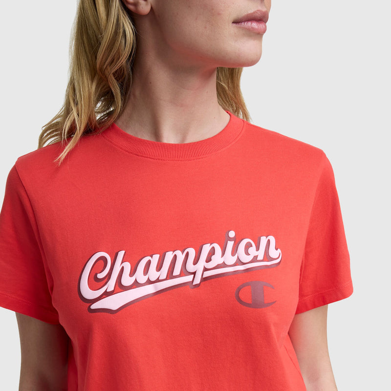 A person wears the Champion Modern Retro Baseball Graphic T-Shirt featuring a white and pink script logo across the chest, against a plain light gray background.