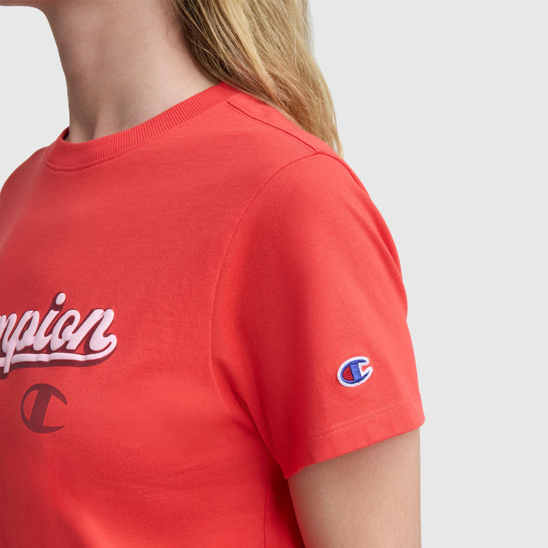 A person wearing a red Champion Modern Retro Baseball Graphic T-Shirt featuring a script logo on the chest and a small embroidered "C" on the left sleeve, against a plain light background.