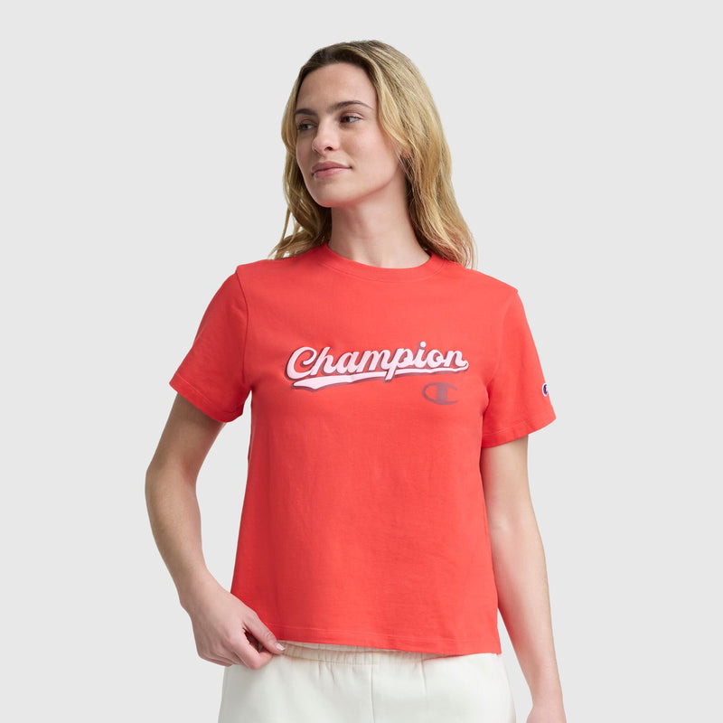 A person with light brown hair wears the Champion Modern Retro Baseball Graphic T-Shirt with a script logo and light pants, standing against a gray background and looking to the side with a relaxed expression.