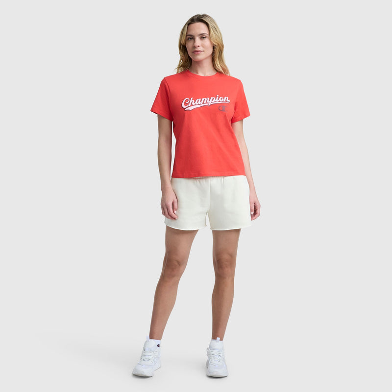 A woman stands facing forward against a plain light grey background, wearing a Champion Modern Retro Baseball Graphic T-Shirt with script logo, paired with white shorts and white sneakers.