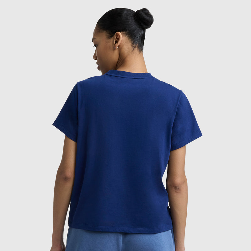 A person with dark hair in a bun is shown from behind, wearing a Champion Summer League Graphic T-Shirt, C Logo in dark blue and light blue pants, standing against a plain light gray background.