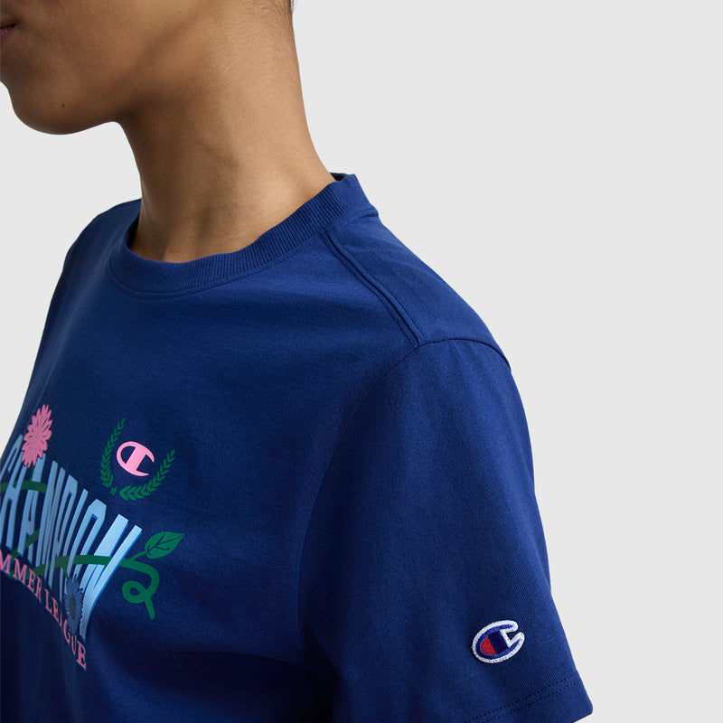 A person wears a royal blue Champion Summer League Graphic T-Shirt with chest floral graphics, C logo patch on the sleeve, and lightweight cotton fabric, shown from neck to upper torso against a plain background.