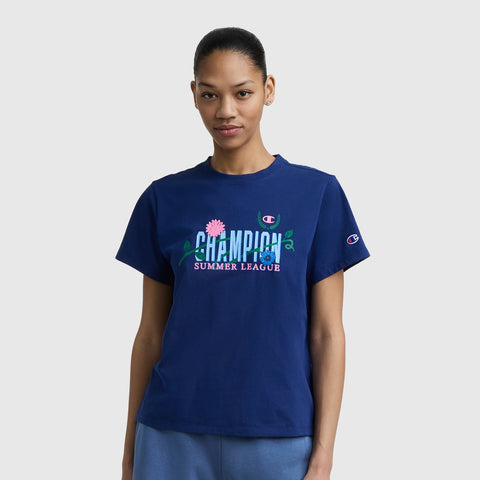 A woman stands against a plain light gray background wearing the Champion Summer League Graphic T-Shirt, featuring a C logo and floral graphics on navy blue lightweight cotton.
