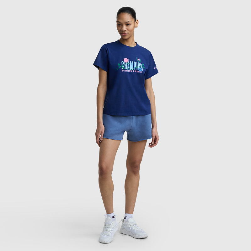 A woman poses against a plain background wearing a Champion Summer League Graphic T-Shirt with a C logo in navy, light blue shorts, white sneakers, and her hair styled in a neat ponytail.