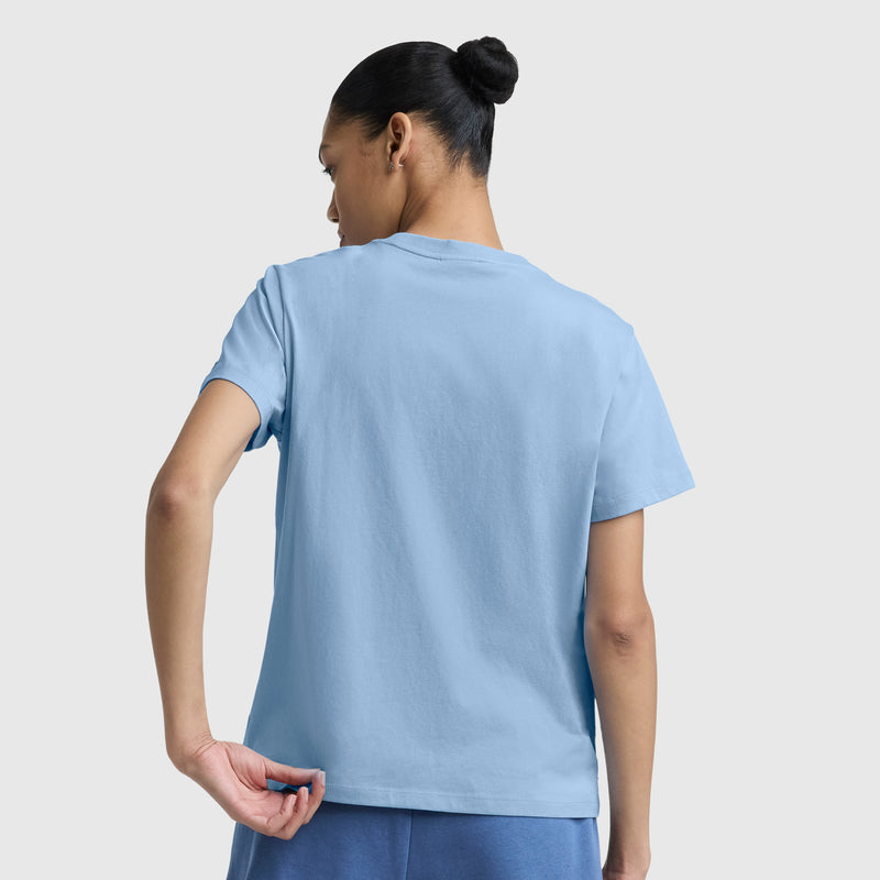 A person with dark hair in a bun is seen from the back, wearing a light blue Champion Blurred Roses Graphic T-Shirt with a C Logo and matching shorts. They hold the hem of their shirt against a plain light background.