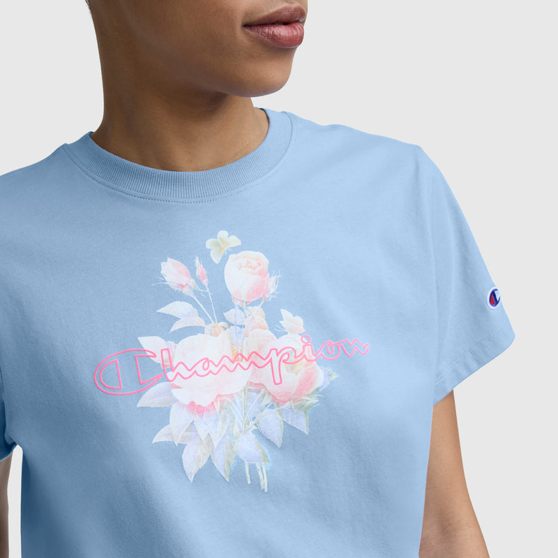 A person wears the Champion Blurred Roses Graphic T-Shirt in light blue combed cotton, featuring a floral design and "Champion" in pink cursive on the front, plus a C logo on the sleeve.