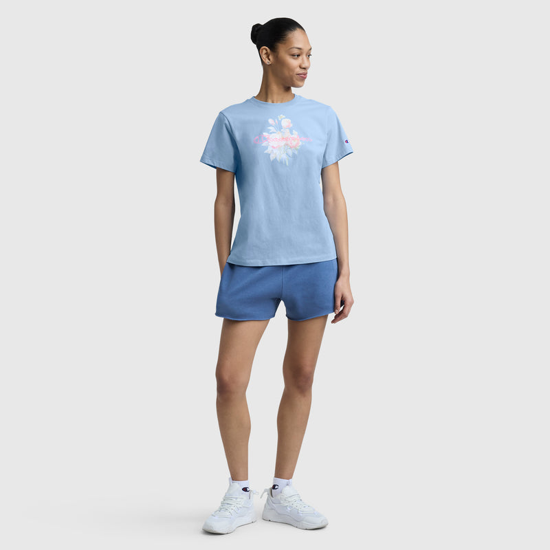 A woman with her hair tied back stands against a plain background wearing a light blue Champion Blurred Roses Graphic T-Shirt with C Logo, blue shorts, and white sneakers, looking to the side with her hands relaxed by her sides.