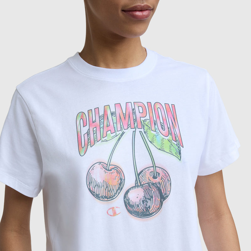 A person wears the Champion Cherry On Top Graphic T-Shirt, a lightweight cotton tee with two cherries, green leaves, bold multicolored "CHAMPION" lettering, and an embroidered C logo on the sleeve.