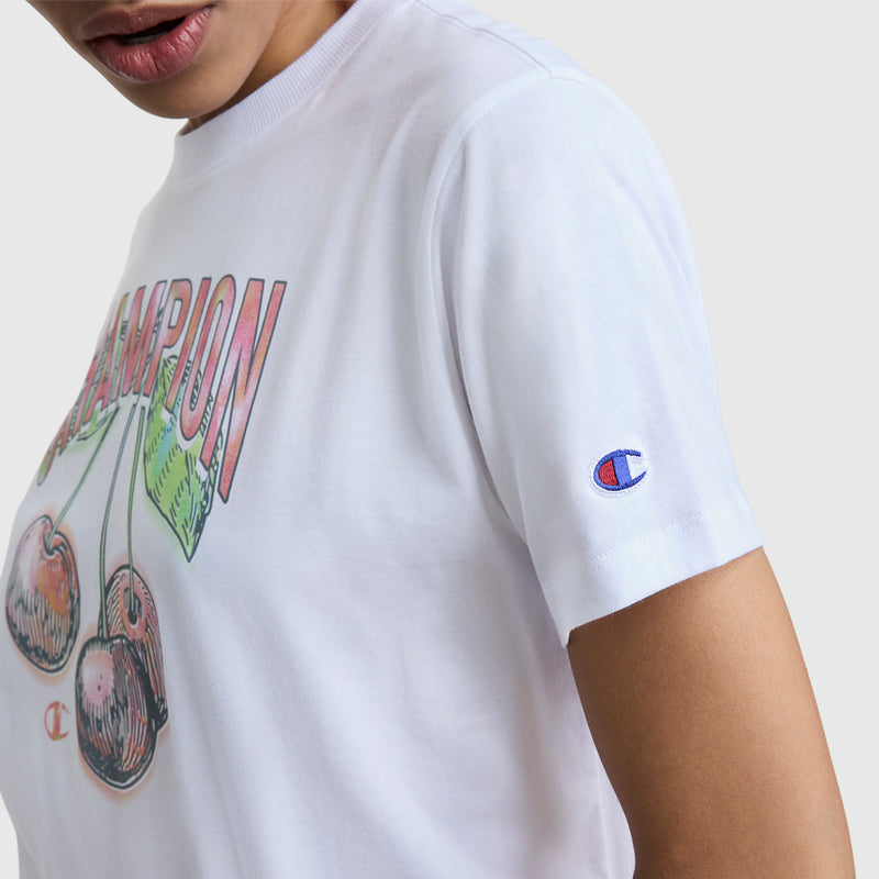 A person is seen wearing a Champion Cherry On Top Graphic T-Shirt in white, featuring an illustrated cherry and CHAMPION text on the front, plus an embroidered C logo patch on the sleeve. Only their lower face and part of their upper body are visible.