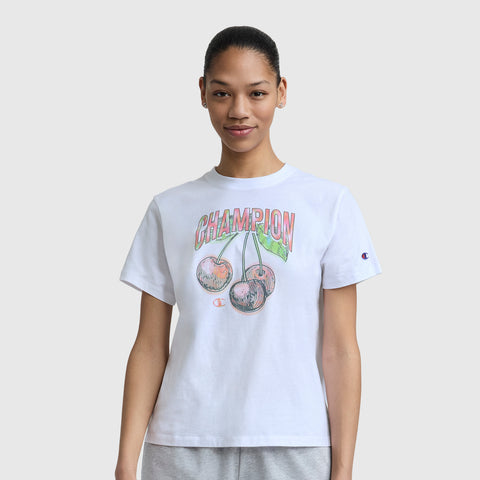 A person wears the Champion Cherry On Top Graphic T-Shirt, featuring CHAMPION text, two illustrated cherries, and an embroidered C logo on lightweight cotton, standing against a plain light gray background.