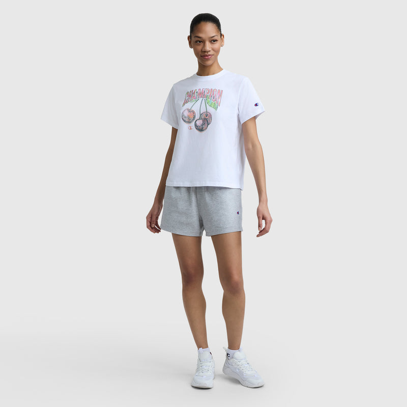 A woman stands against a light gray background, smiling softly with her hair tied back. She wears a Champion Cherry On Top Graphic T-Shirt (C Logo), light gray shorts, and white sneakers.