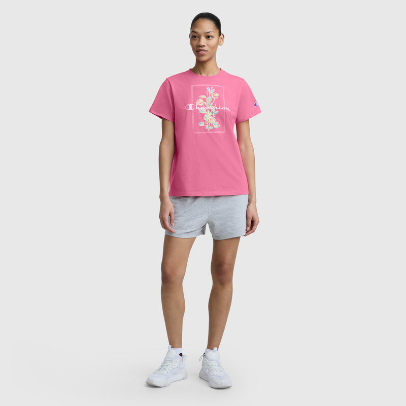 A woman stands facing forward, wearing a pink Champion Painted Bouquet Graphic T-Shirt with C Logo, paired with gray shorts and white sneakers, against a plain white background.