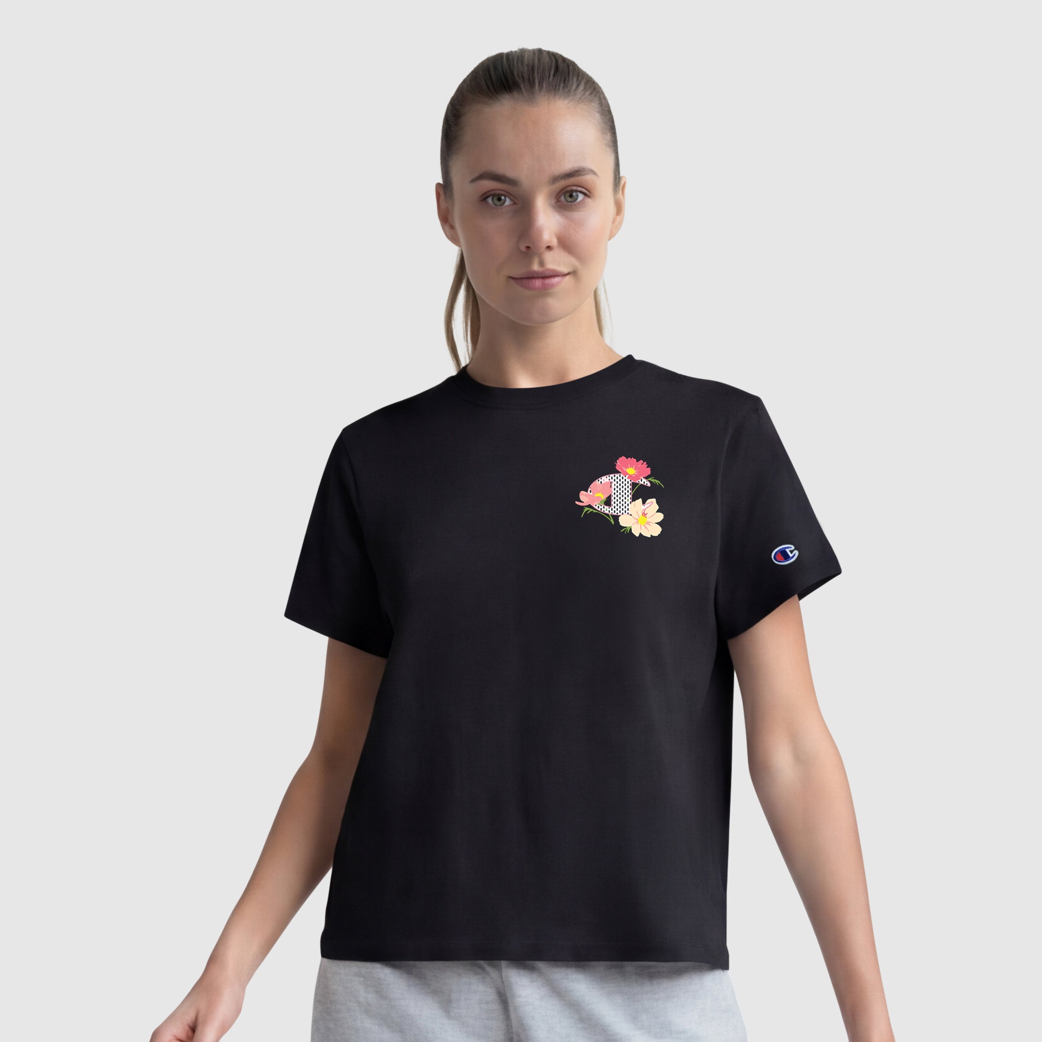 A woman wears a black Champion Floral Mesh "C" Graphic T-Shirt featuring a small mesh C logo on the left chest and gray shorts, standing against a plain light gray background.