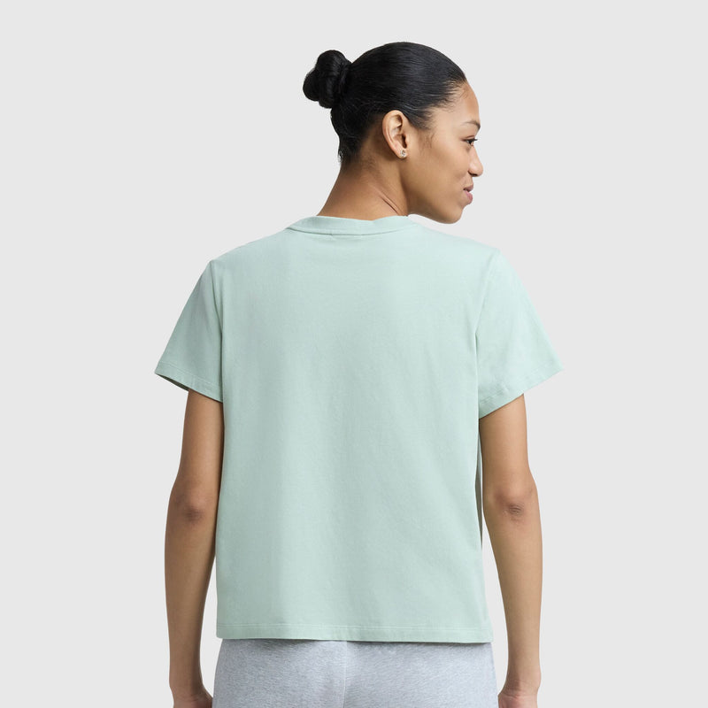 A woman with dark hair in a bun is seen from behind, wearing a Champion Floral Mesh "C" Graphic T-Shirt and gray pants against a plain light background.