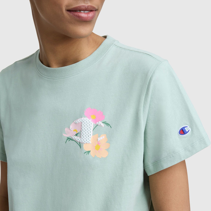 A person is shown from the shoulders up wearing a Champion Floral Mesh "C" Graphic T-Shirt with a logo on the chest and sleeve, set against a plain background.
