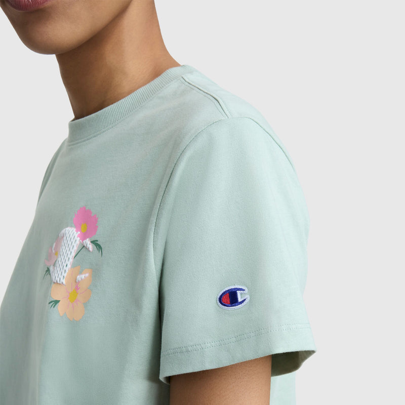 A person wears a light green Champion Floral Mesh "C" Graphic T-Shirt with a floral chest design and C logo patch on the sleeve, shown from neck to upper arm against a plain background.
