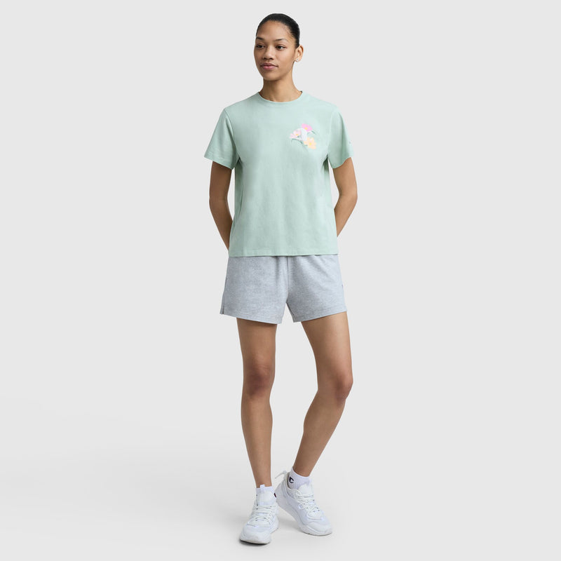 A woman poses against a plain background wearing the Champion Floral Mesh "C" Graphic T-Shirt and gray shorts, with her hair tied back and one hand on her thigh. White sneakers complete her casual look.