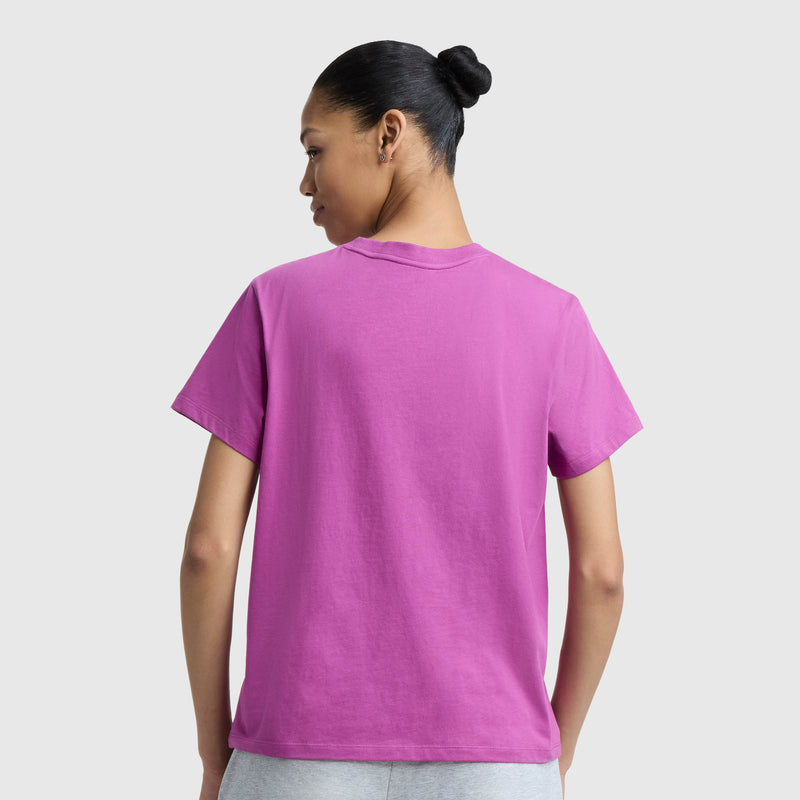 A woman with her hair in a bun stands with her back to the camera, wearing a bright purple Champion Butterfly Collage Graphic T-Shirt with a C Logo and light gray pants against a plain light background.