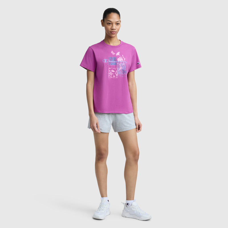 A woman faces forward against a plain light background, wearing a bright pink Champion Butterfly Collage Graphic T-Shirt with C Logo, light gray shorts, and white athletic shoes.