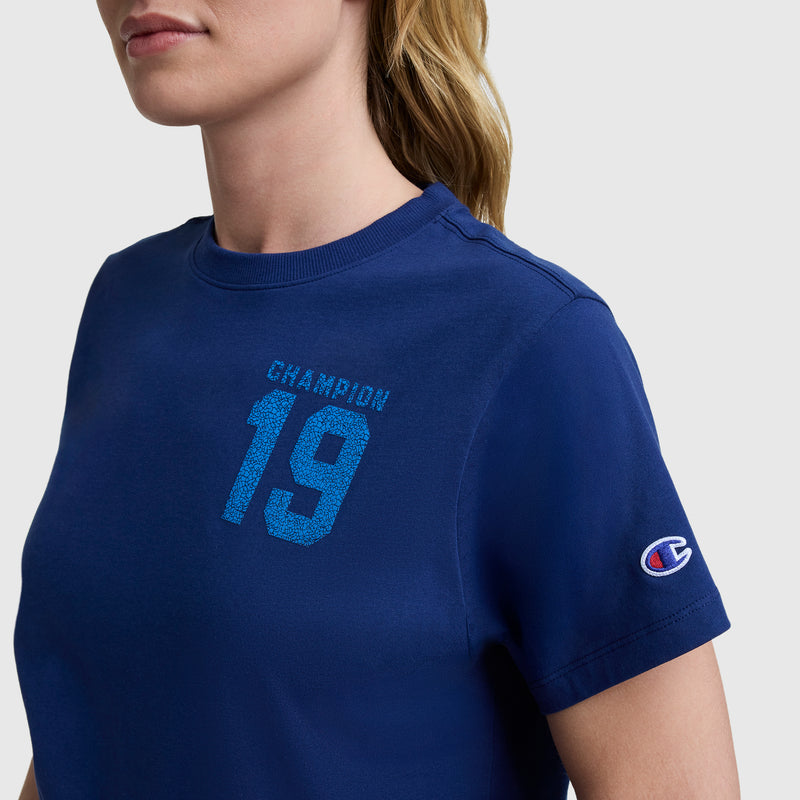 A woman wears the Champion Grid Iron '19 Graphic T-Shirt with "CHAMPION 19" on the chest and a C logo on the sleeve. Only her upper torso and lower face are visible, highlighting a sporty style.