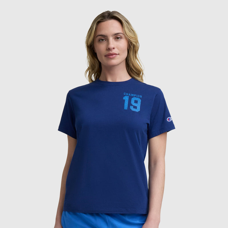 A woman with wavy blonde hair wears the Champion Grid Iron '19 Graphic T-Shirt with a C logo, featuring a navy blue design, standing against a plain light gray background.