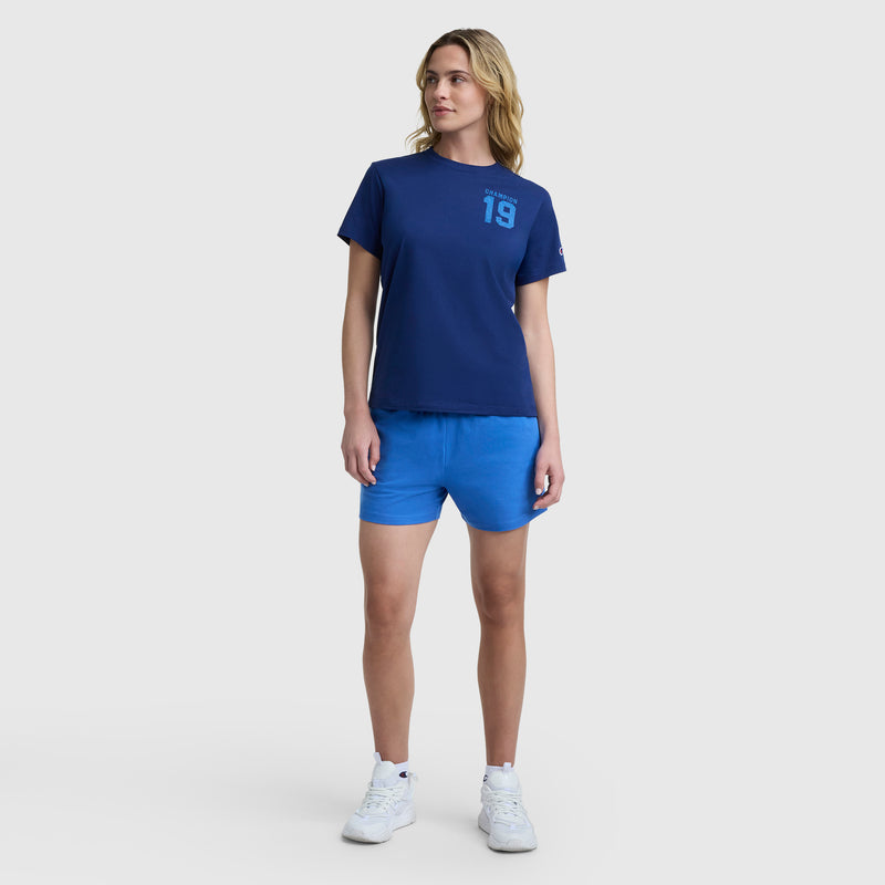 A woman with blonde, wavy hair stands facing forward, wearing the Champion Grid Iron '19 Graphic T-Shirt with a C logo, bright blue shorts, and white sneakers. She looks slightly to her left in this vintage athletic style.