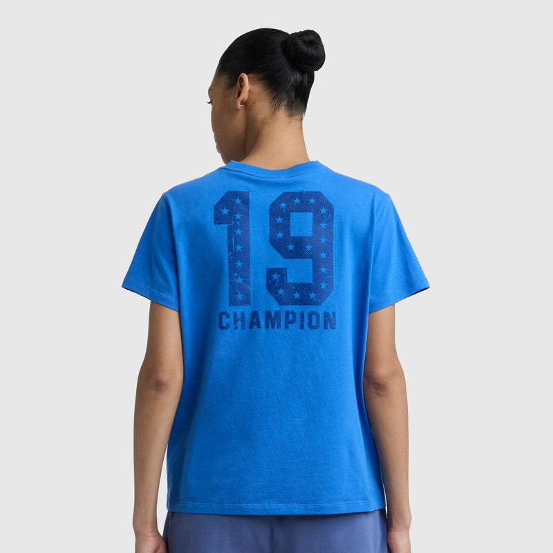 A person with hair in a bun wears the Champion Grid Iron '19 Graphic T-Shirt featuring the C logo and large "19" and "CHAMPION" print on the back, styled sportily against a plain light background.